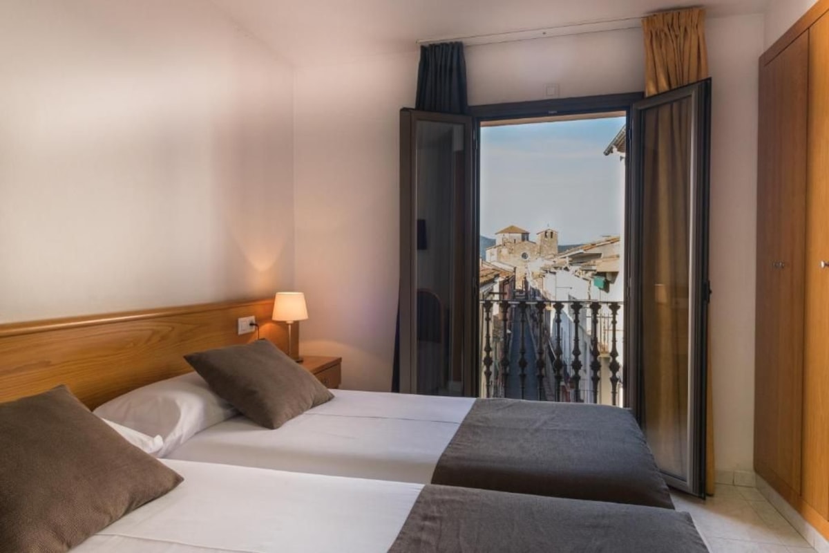 A hotel room with a bed, a nightstand, a lamp, and a view of a cityscape through an open door.