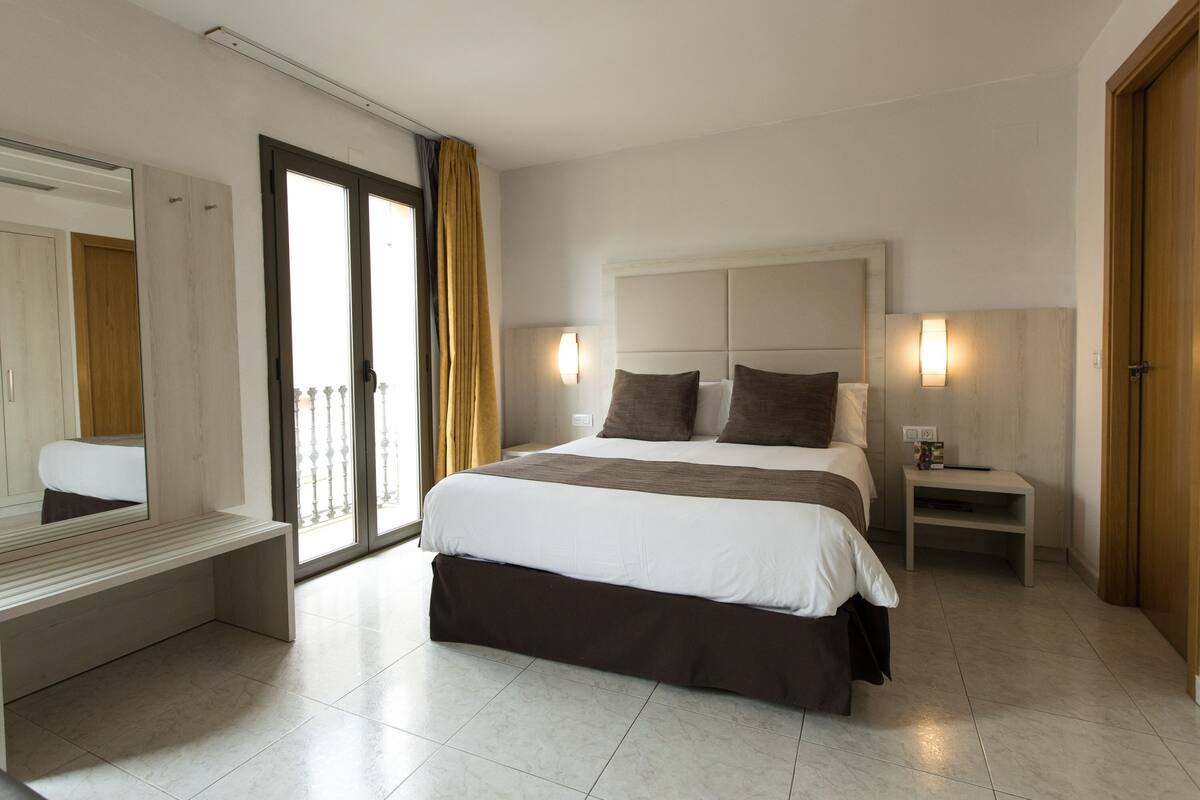 Una habitación de hotel moderna con una cama grande, un banco, una mesita de noche y un espejo.