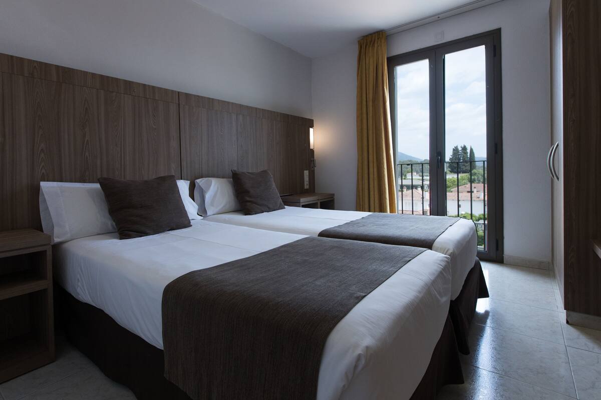 Habitación de hotel con dos camas, cabeceros de madera, mesitas de noche y un balcón con vistas.