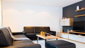 Een moderne woonkamer met een zwarte leren bank, een houten salontafel en een zwart poefje. Er is een flatscreen televisie op een houten standaard en een klein keukenhoekje met kastjes en een magnetron.