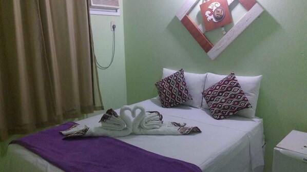 A hotel room with a bed, pillows, a towel arrangement, a window with curtains, and a wall-mounted air conditioning unit.