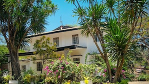 A two-story building with a white facade and dark roof, surrounded by lush greenery and flowering plants. - Lac s Villa (Freetown)