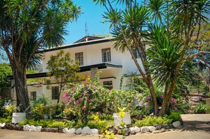 A two-story building with a white facade and dark roof, surrounded by lush greenery and flowering plants.
