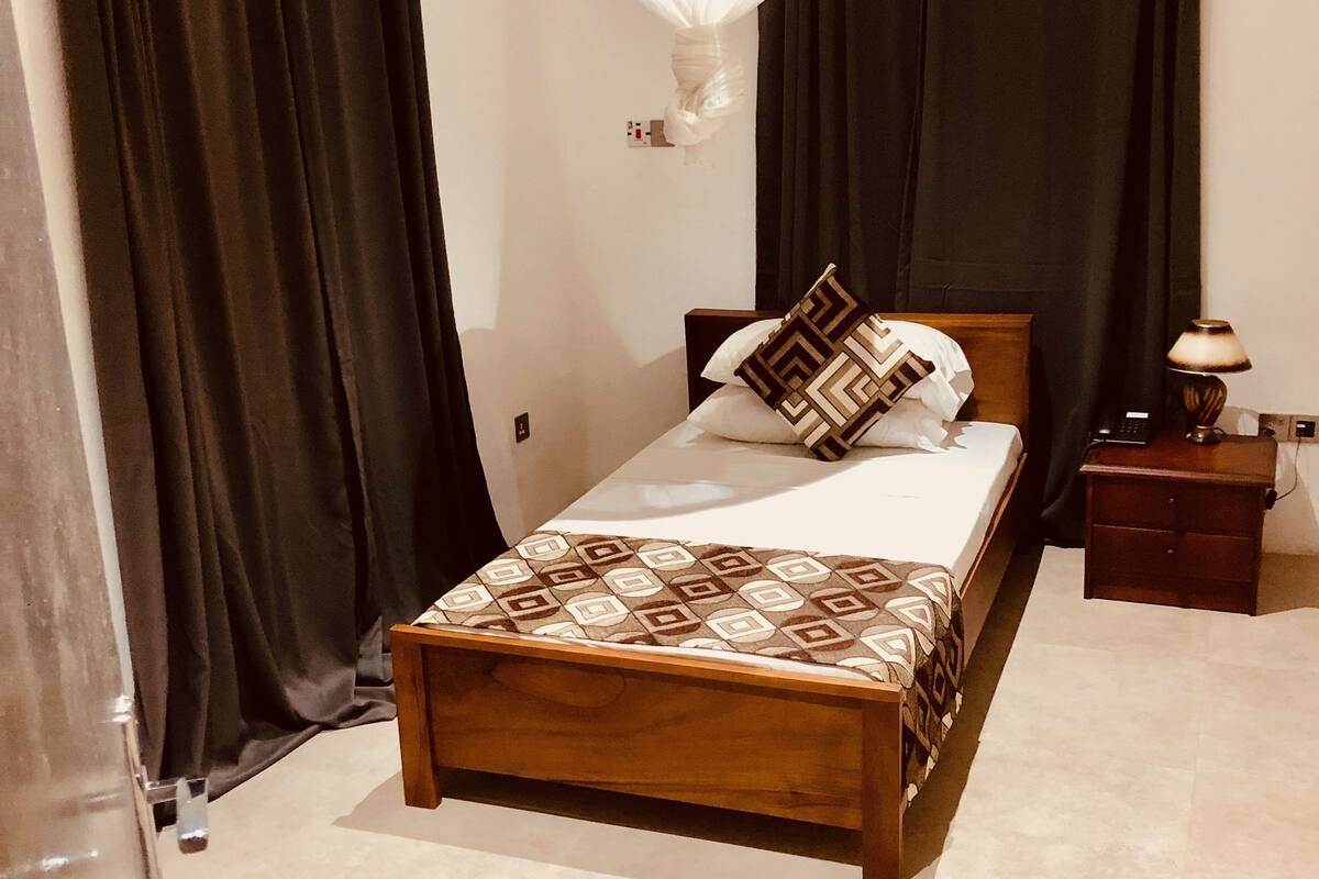 A bedroom with a wooden bed, black curtains, a white hanging light fixture, a bedside table with a lamp, and a tiled floor.