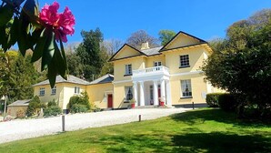 A large, yellow building with a white balcony, surrounded by greenery and a clear blue sky.