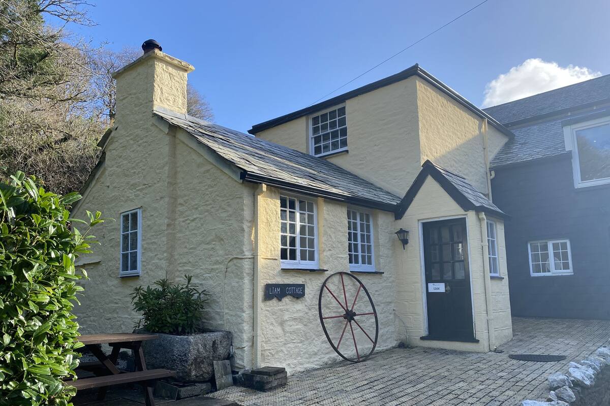 A traditional stone cottage with a large wheel, a wooden bench, and a sign reading "Liam Cottage."