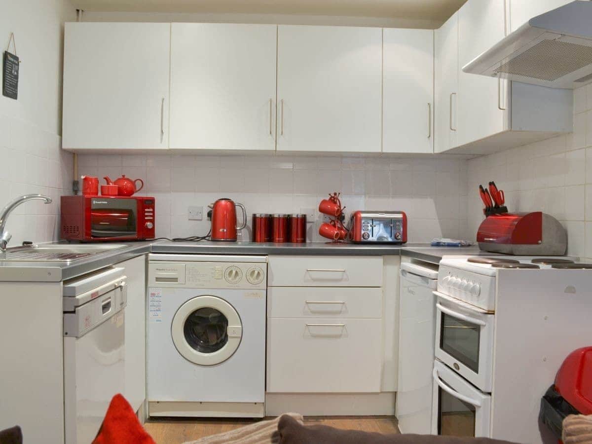 Een compacte keuken met witte kastjes, een wasmachine, een magnetron, een broodrooster en een rode waterkoker.