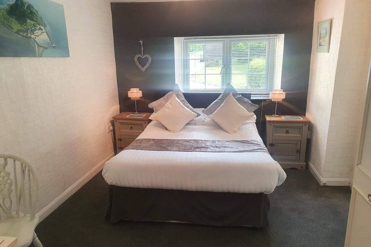 A bedroom with a large bed, two bedside tables, a window with blinds, and a wall-mounted heart decoration.