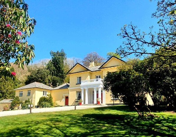 Un edificio grande y amarillo con un porche blanco y columnas, rodeado de abundante follaje y árboles.