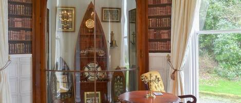 A room with a large arched doorway leading to a library filled with bookshelves. There is a round wooden table with a brass candelabra and a yellow upholstered chair.