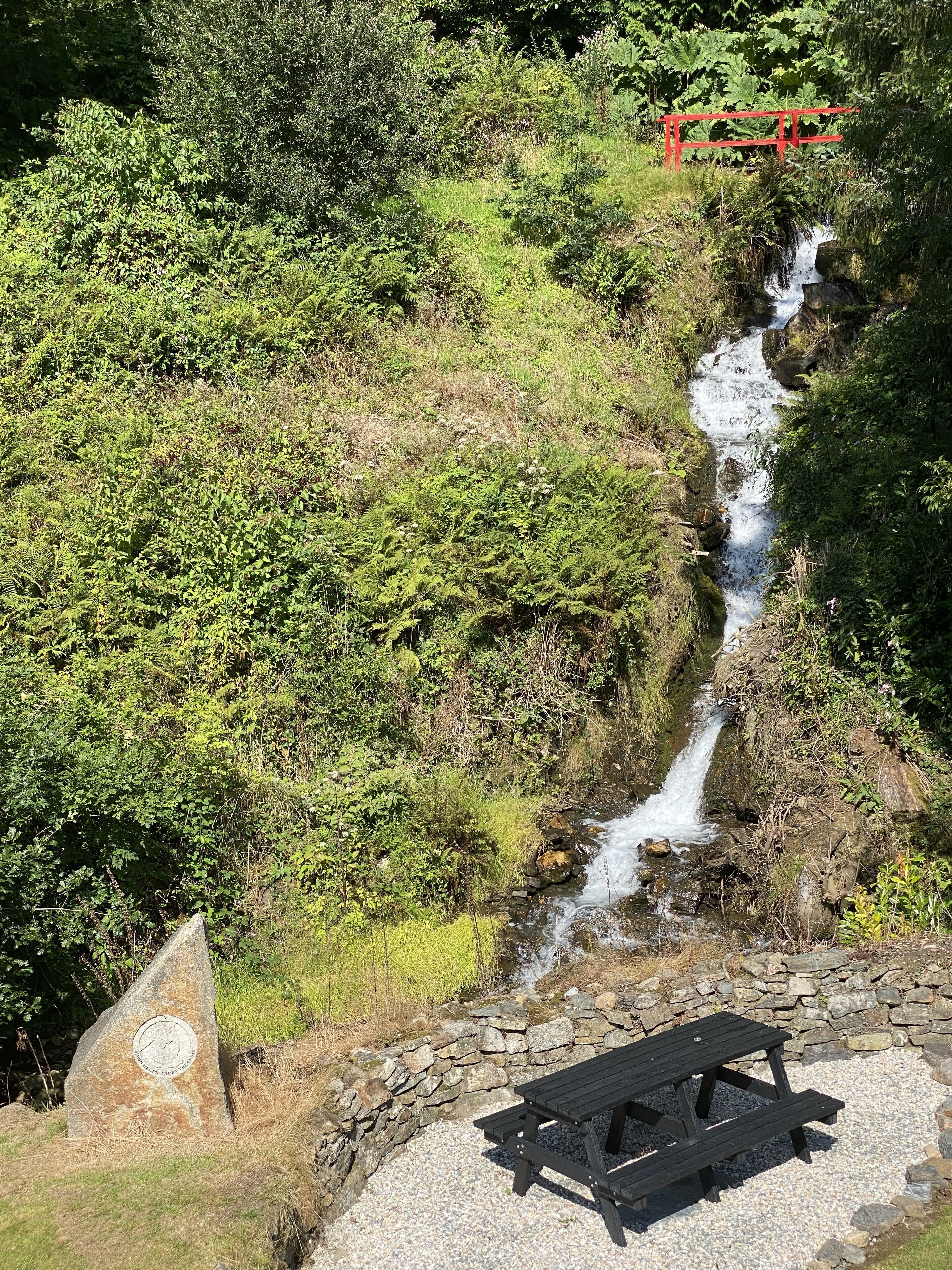 A picnic table and a stone monument in a natural setting with a waterfall and dense greenery.