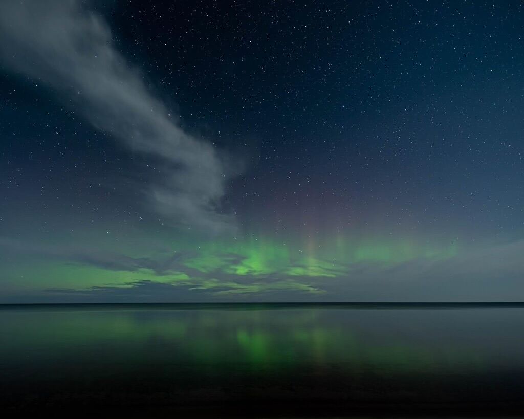 Cielo nocturno con estrellas y la aurora boreal sobre una superficie acuática tranquila.
