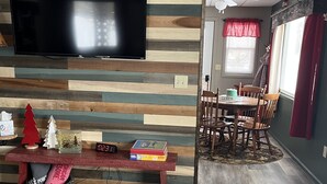 Una sala de estar con una televisión instalada en una pared de madera, una zona de comedor con una mesa y sillas, y una alfombra estampada en el suelo.