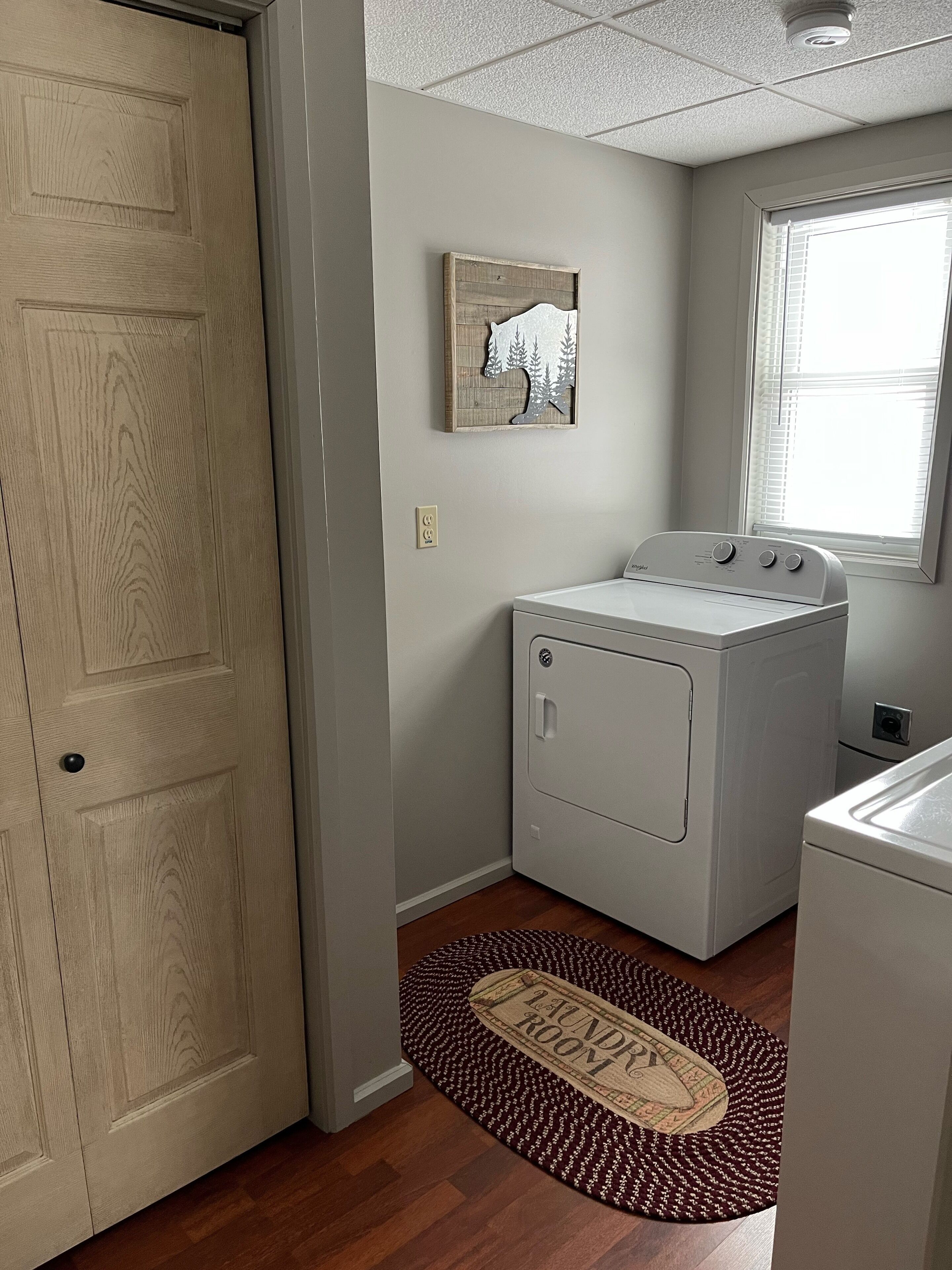 A laundry room with a washer and dryer, a wooden door, a wall-mounted artwork, and a window with blinds.