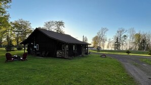 Una cabaña rústica con un porche y dos sillas Adirondack, rodeada de un campo cubierto de hierba y árboles.