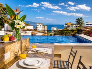 Ein Essbereich am Pool mit Blick auf das Meer, ein Tisch gedeckt mit Tellern und einem Glas Orangensaft, sowie ein Stuhl.