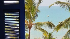 Una habitación con vista al océano, que incluye una cama con cubrecama azul de punto, una ventana con persianas horizontales y una palmera en el fondo.