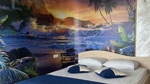 A bedroom with a large mural of a sunset over the ocean, a bed with blue bedding, and a nightstand. - Kirch Hotel & Restaurant (Goris)