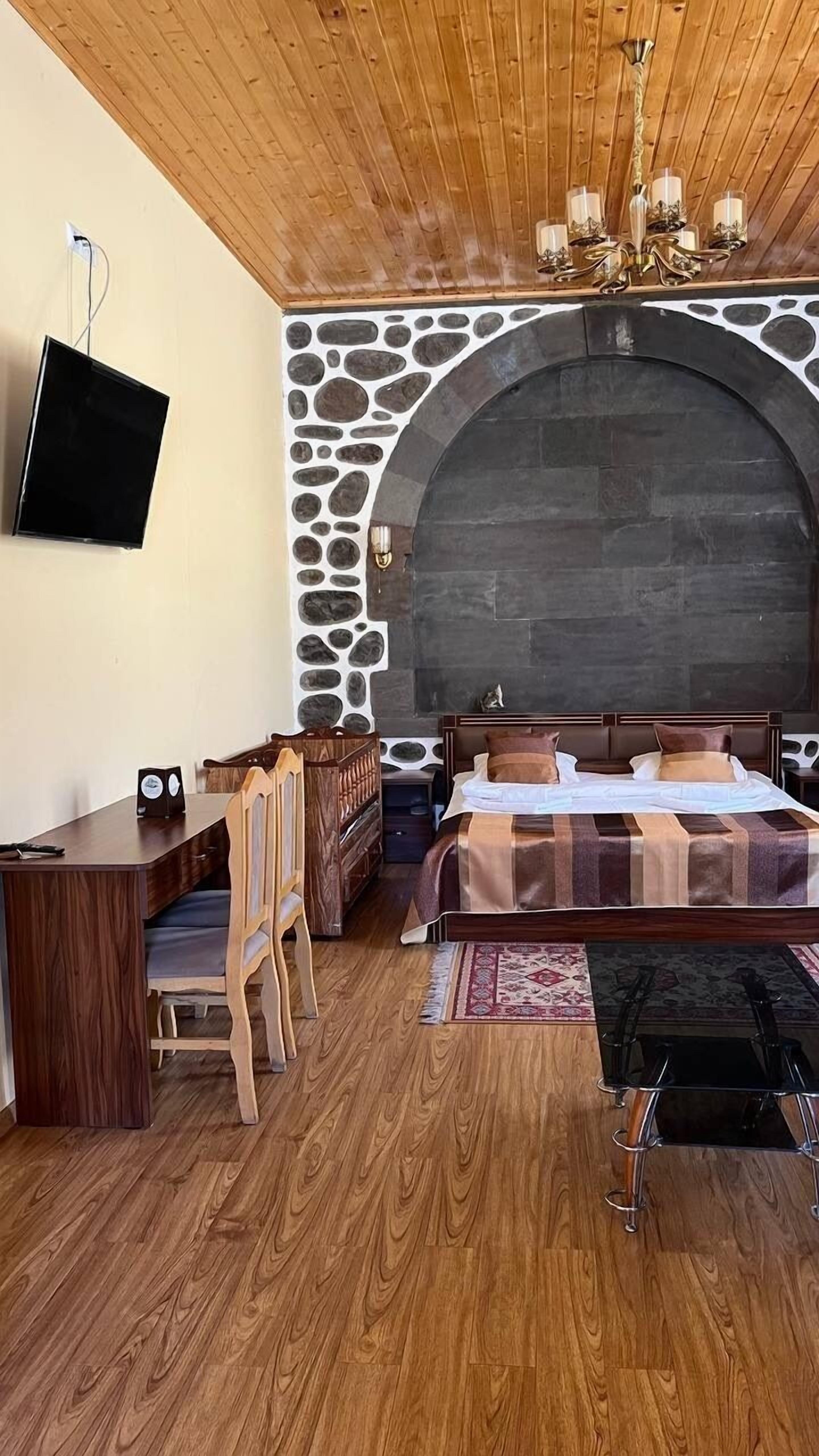 Une chambre avec un mur en pierre, un plafond en bois, un lit, un bureau et un téléviseur fixé au mur.