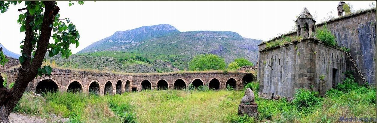 An old stone bridge with multiple arches, a rustic building with a tower, and a mountainous backdrop.