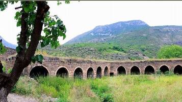 An old stone bridge with multiple arches, a rustic building with a tower, and a mountainous backdrop.