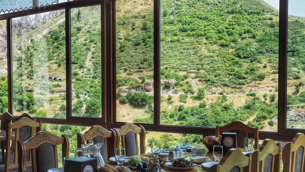 A dining table set with plates, glasses, and a speaker, overlooking a mountainous landscape. - Kirch Hotel & Restaurant (Goris)
