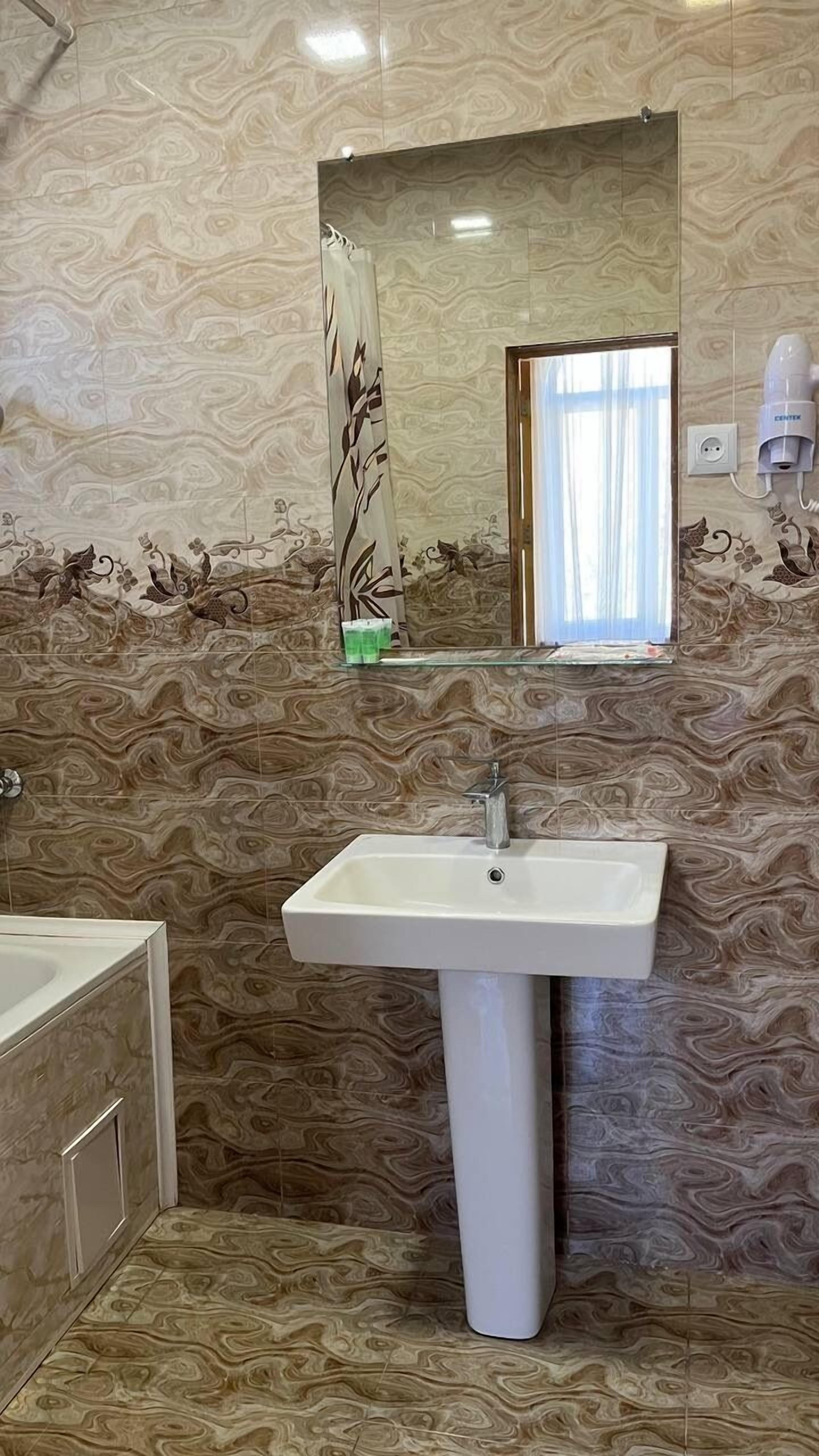 Un baño con pared de diseño de mármol, un lavamanos pedestal blanco, una repisa de vidrio y un secador de pelo en la pared.