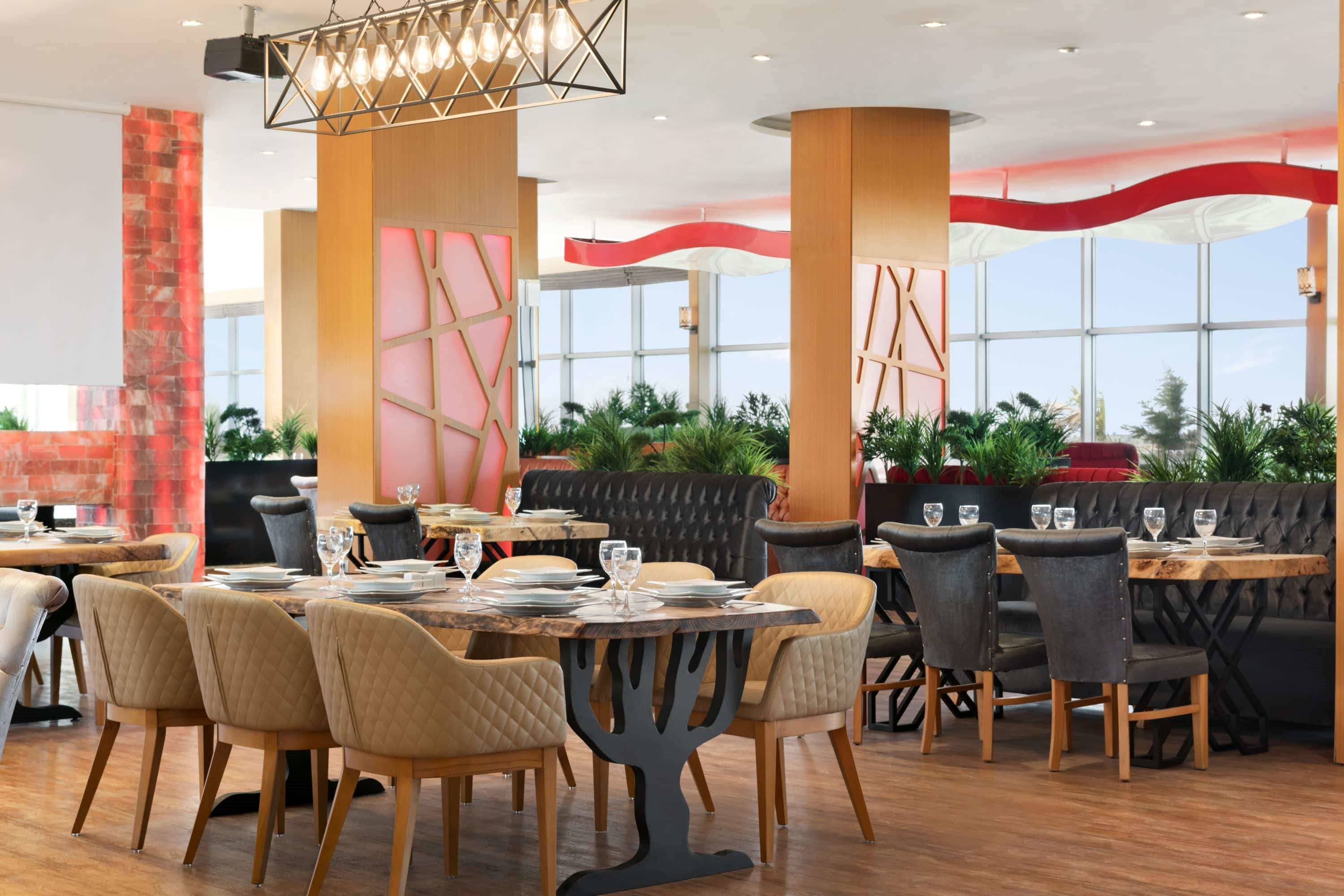 A modern restaurant with wooden tables and chairs, large windows, and potted plants.