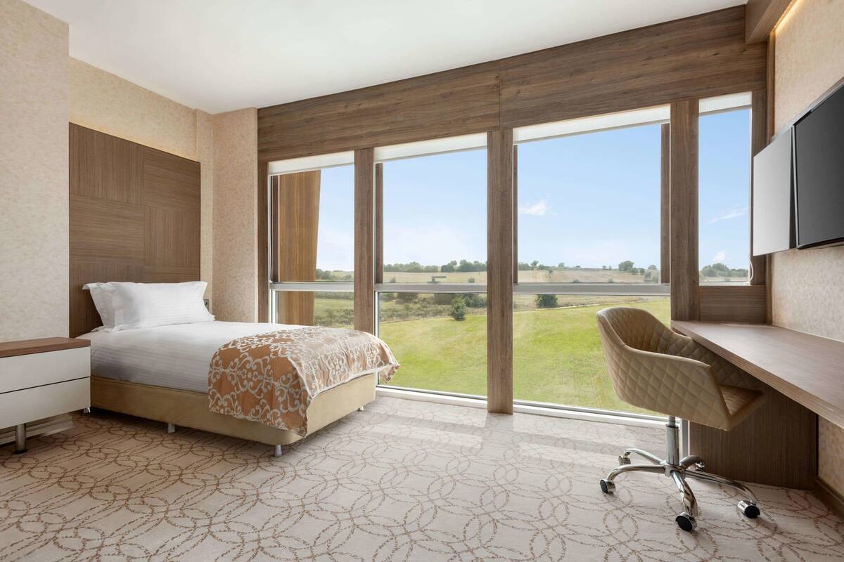A modern hotel room with a large bed, a desk, and a view of a green landscape through large windows.