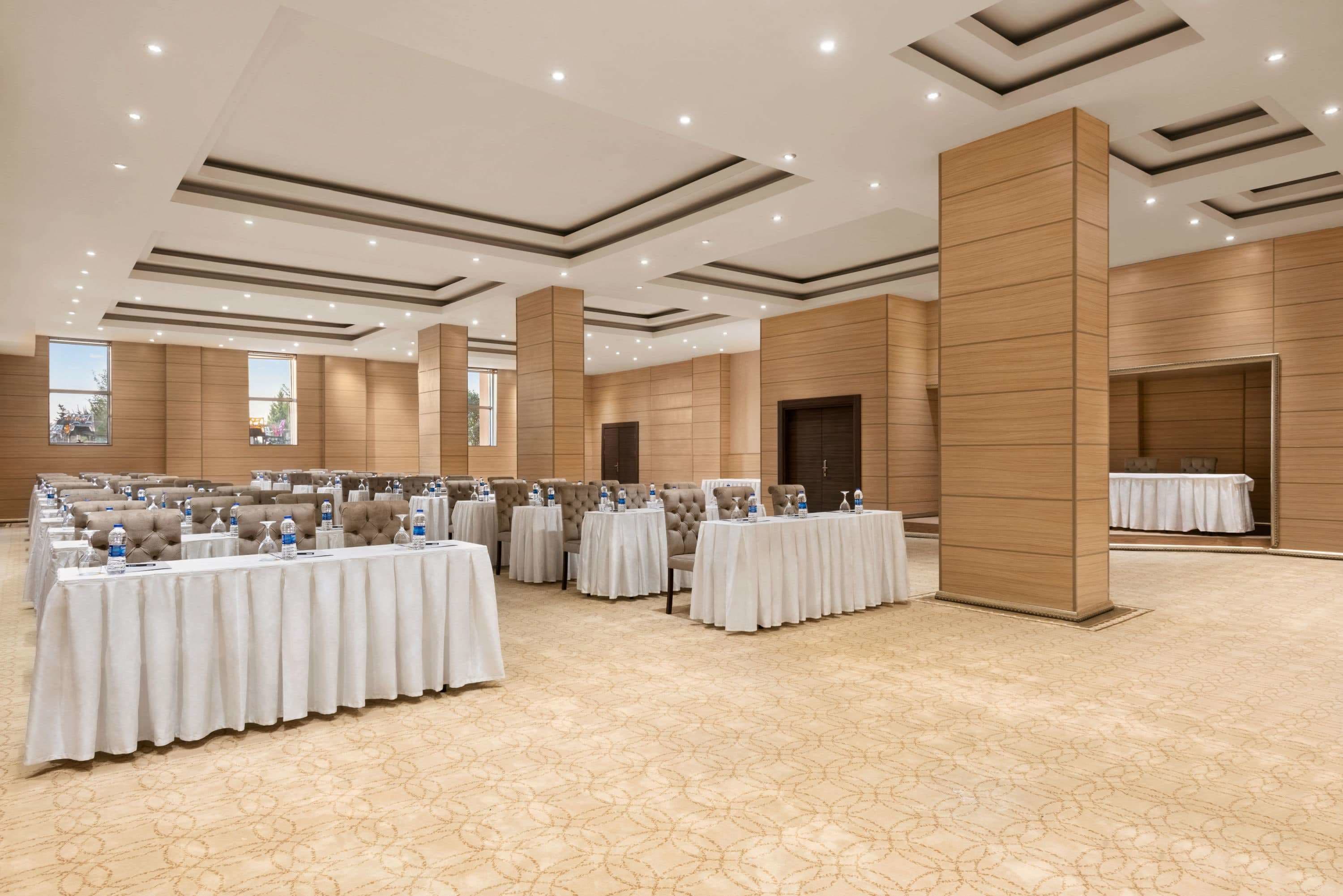 Una amplia sala de conferencias con varias mesas, sillas y botellas de agua, que cuenta con un diseño de techo moderno y ventanas grandes.