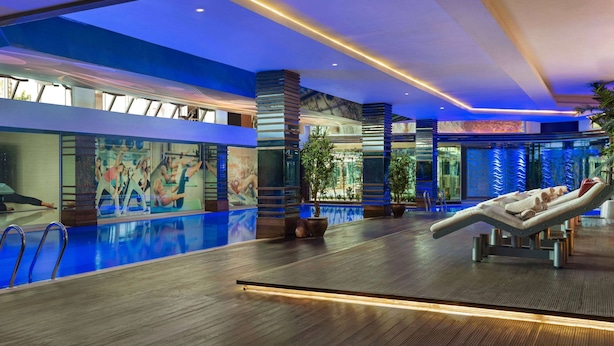 A modern indoor pool area with a glass wall, a lounge chair, and a ceiling with blue lighting.