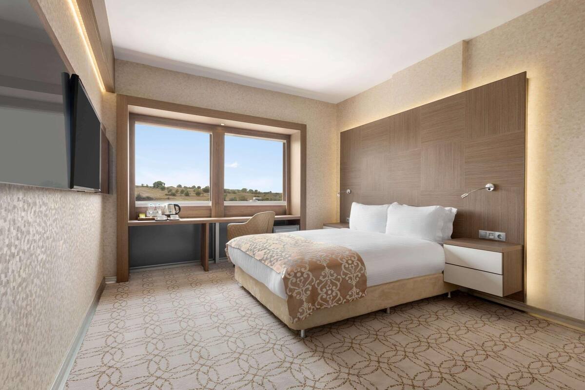 A modern hotel room with a large bed, a desk with a chair, a bedside table, and a view of the outdoors.