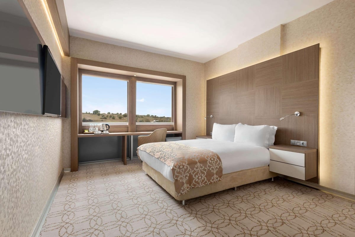 A modern hotel room with a large bed, a desk with a chair, a bedside table, and a view of the outdoors.