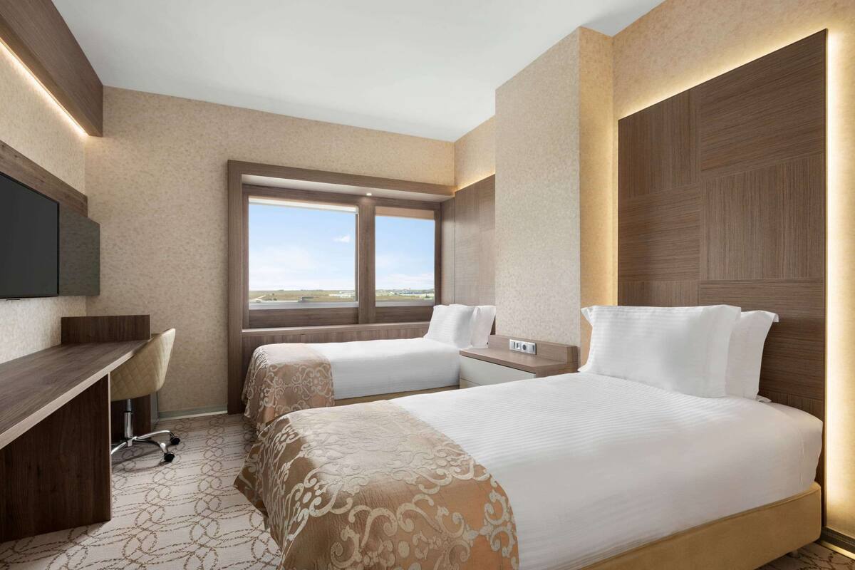 A hotel room with two beds, a desk, a chair, and a large window offering a view.