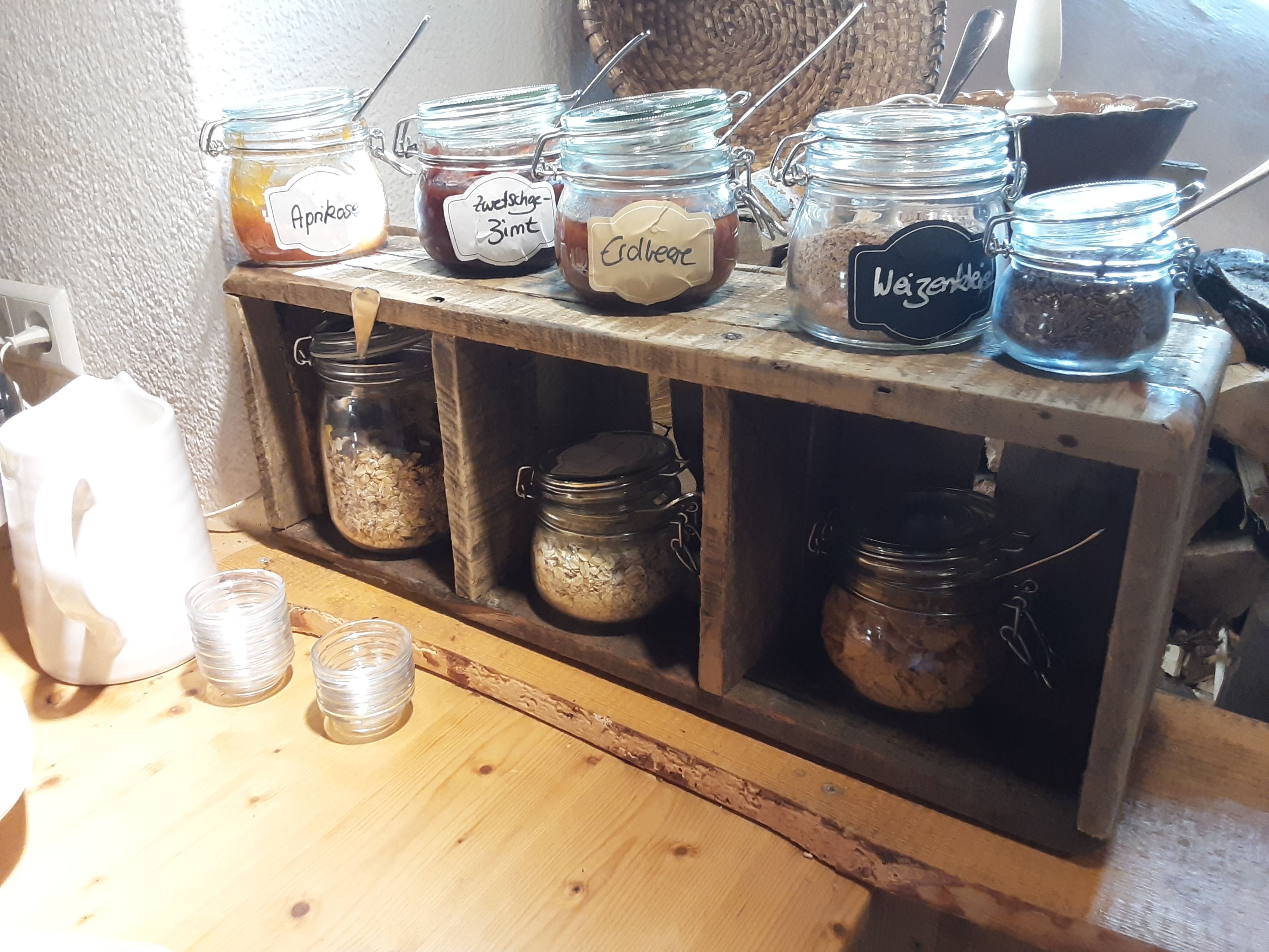 Een houten plank met meerdere glazen potten, voorzien van etiketten met inhoud zoals abrikozen, chocolade en koffie.