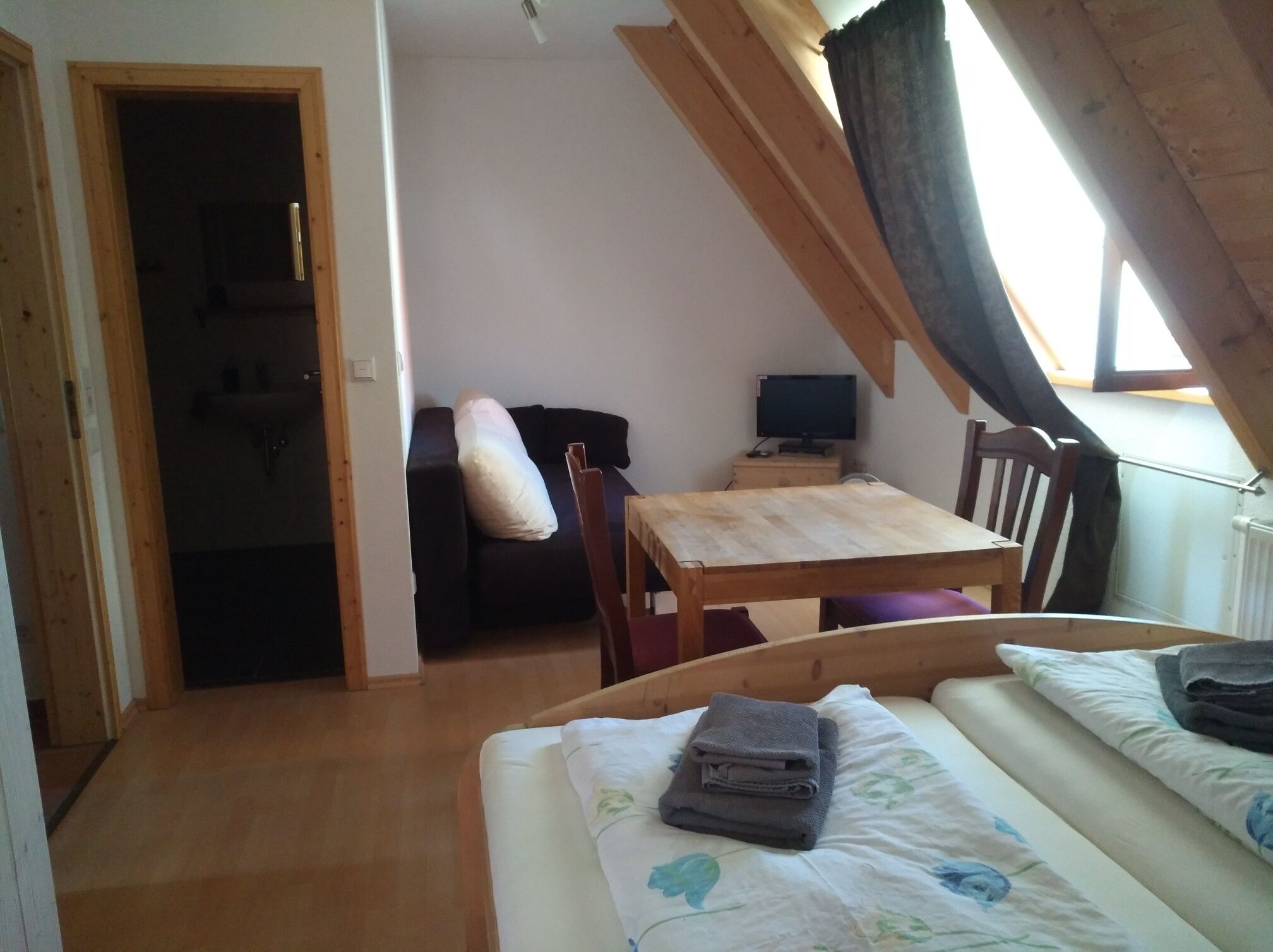 A small, cozy room with a bed, a wooden table, a chair, and a desk with a computer.