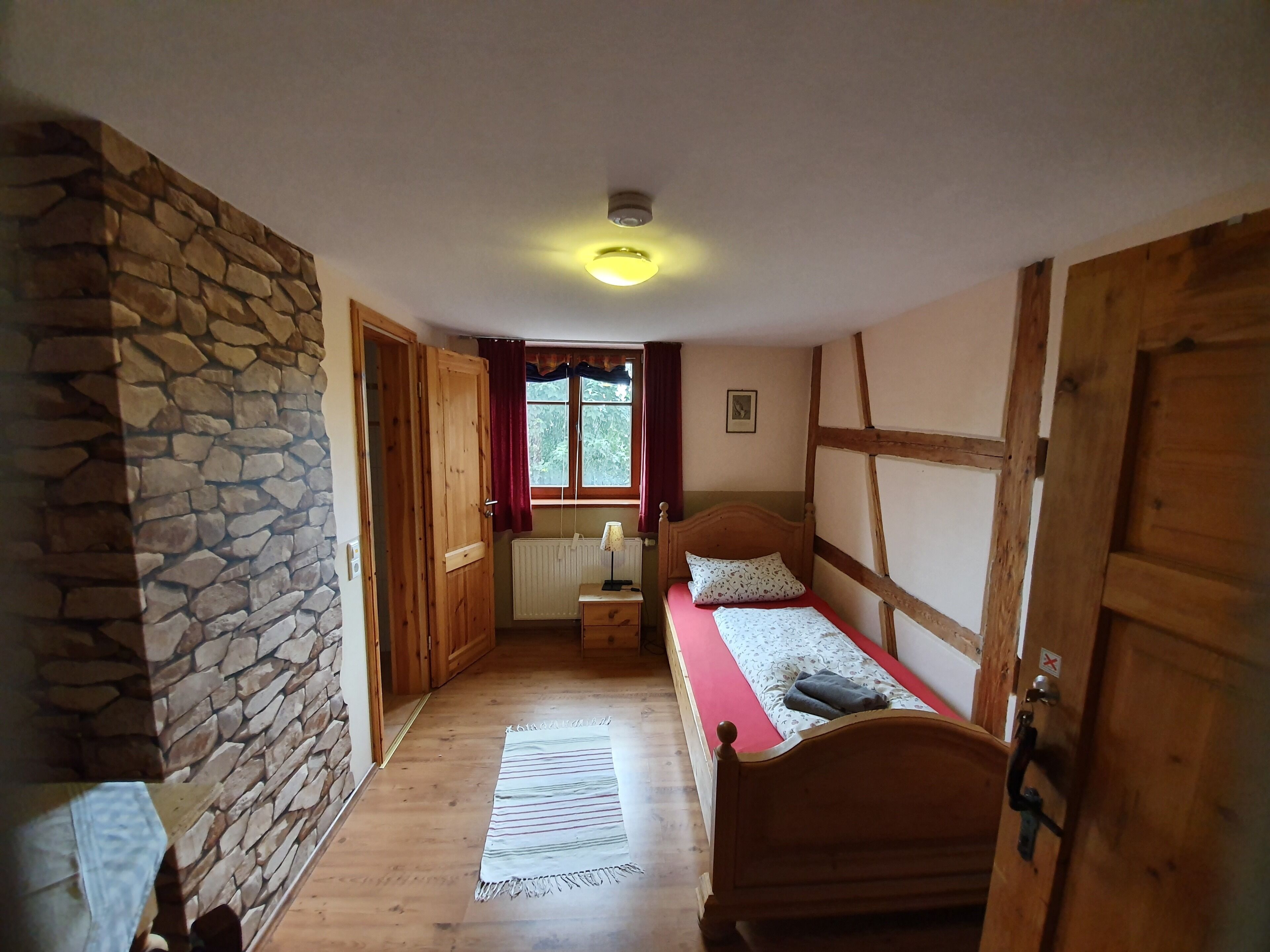 Un pasillo estrecho que conduce a un dormitorio con una cama de madera, pared de piedra y una ventana con cortinas rojas.