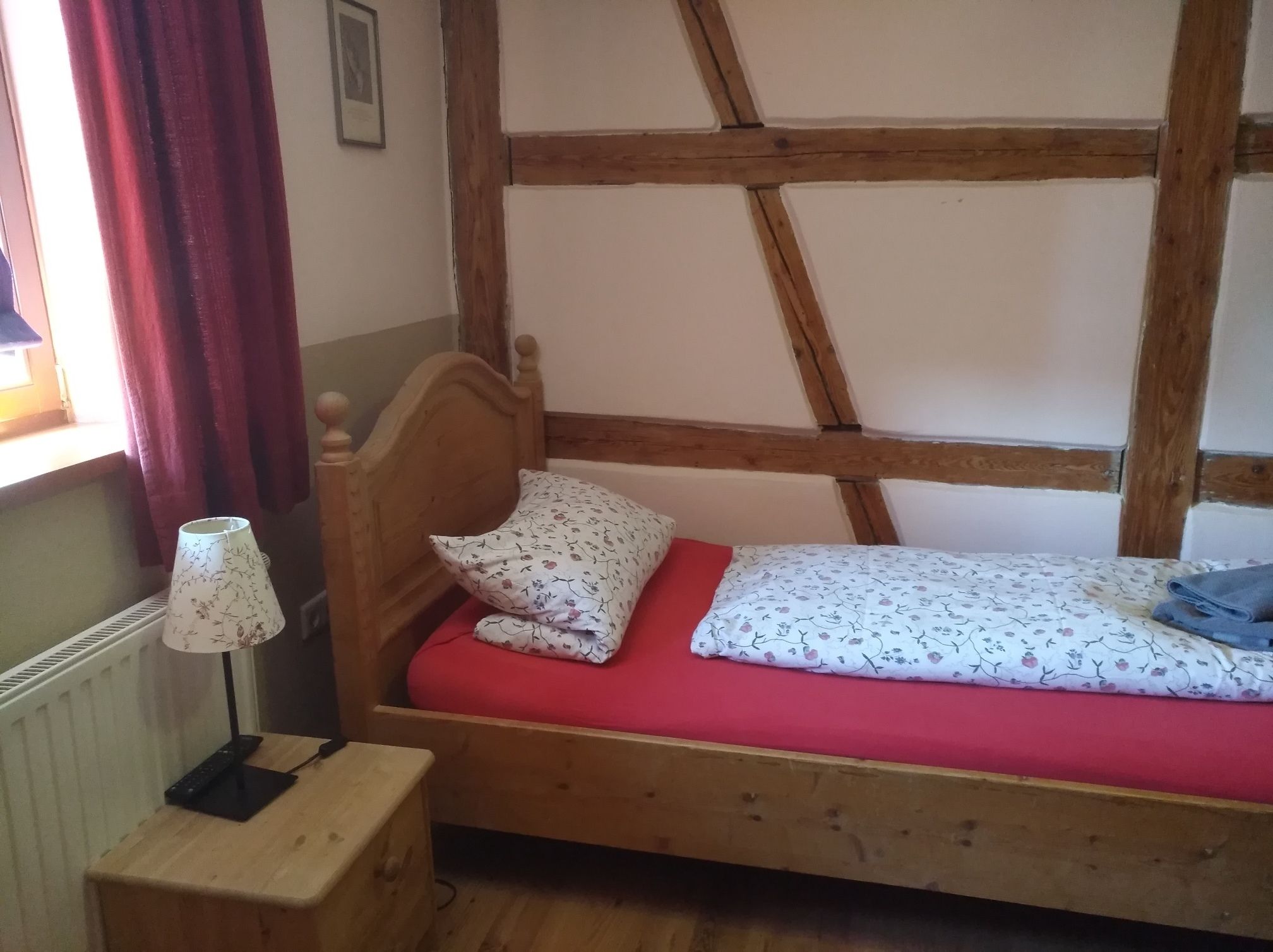 A small, single-bed room with a wooden headboard, a red mattress, and a bedside table with a lamp.