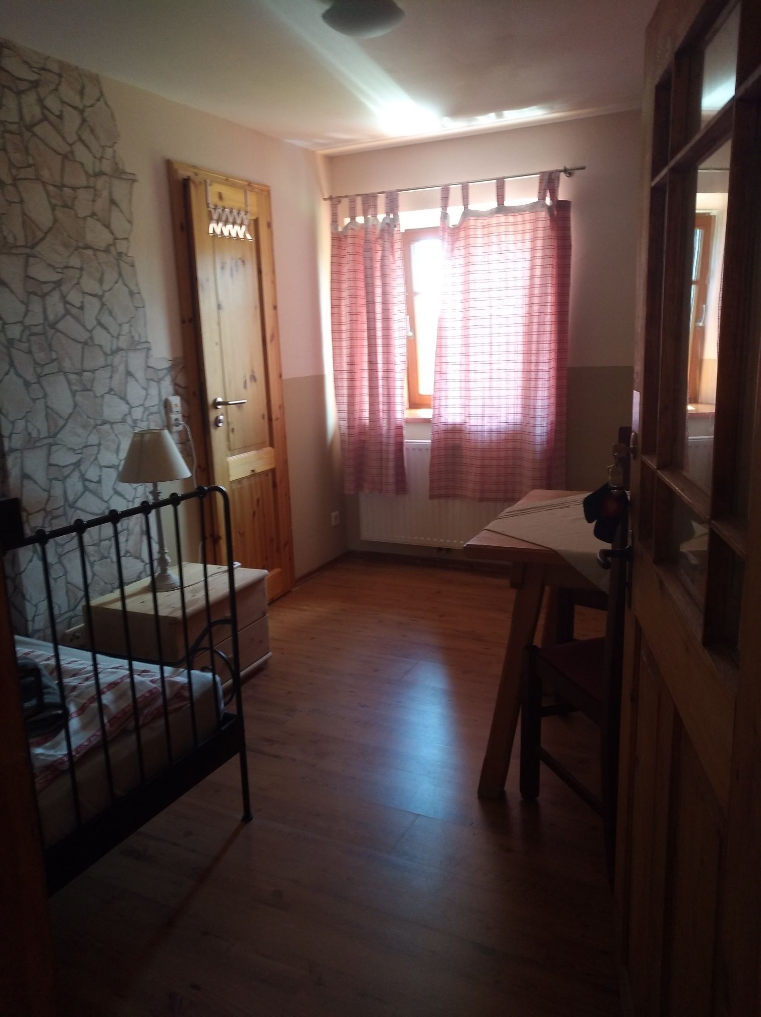 Una habitación con pared de piedra, cama con estructura de metal, puerta de madera, ventana con cortinas y un radiador.
