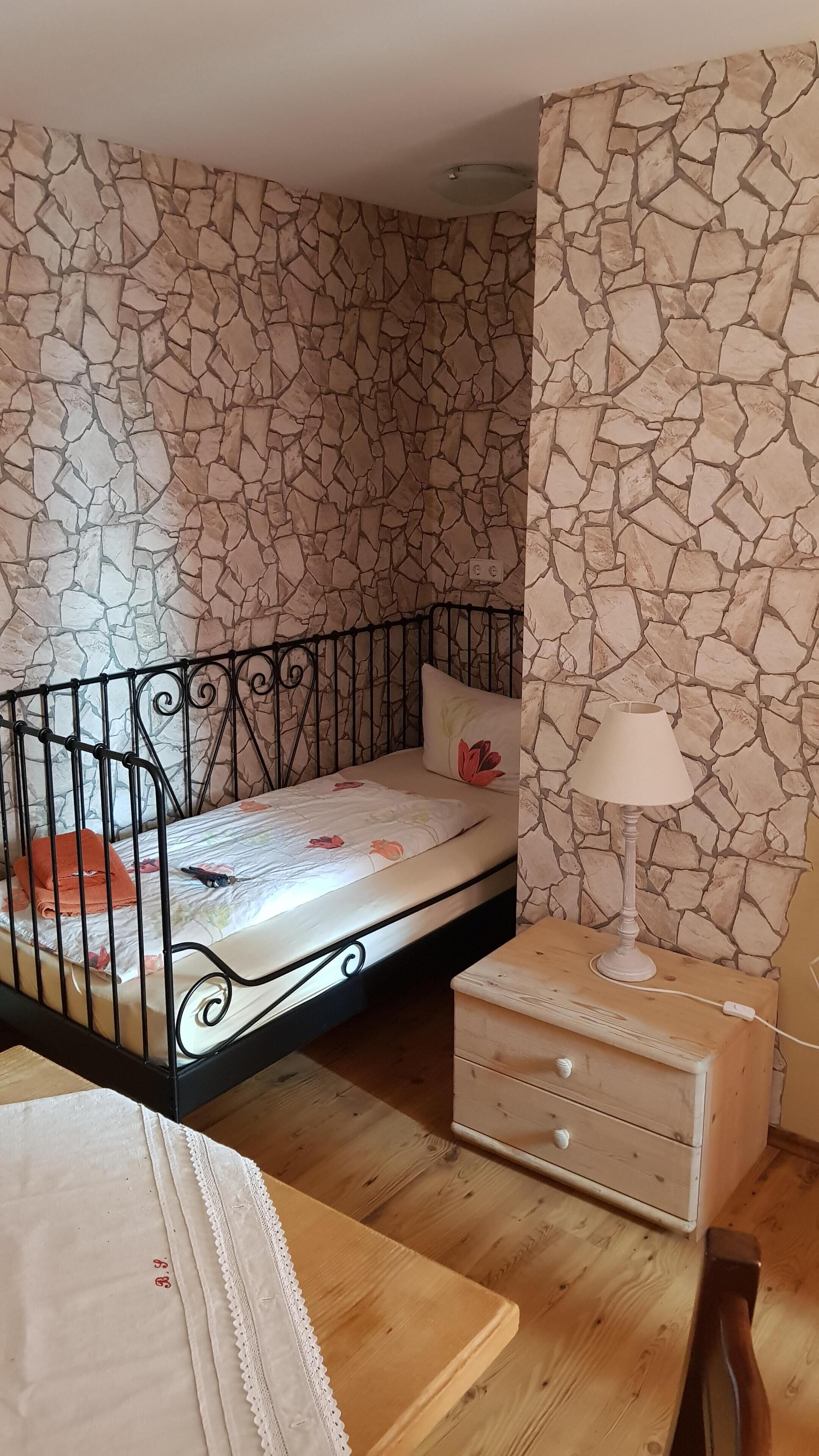 Un dormitorio con pared de diseño de piedra, estructura de cama metálica, mesita de noche con lámpara y cómoda de madera.