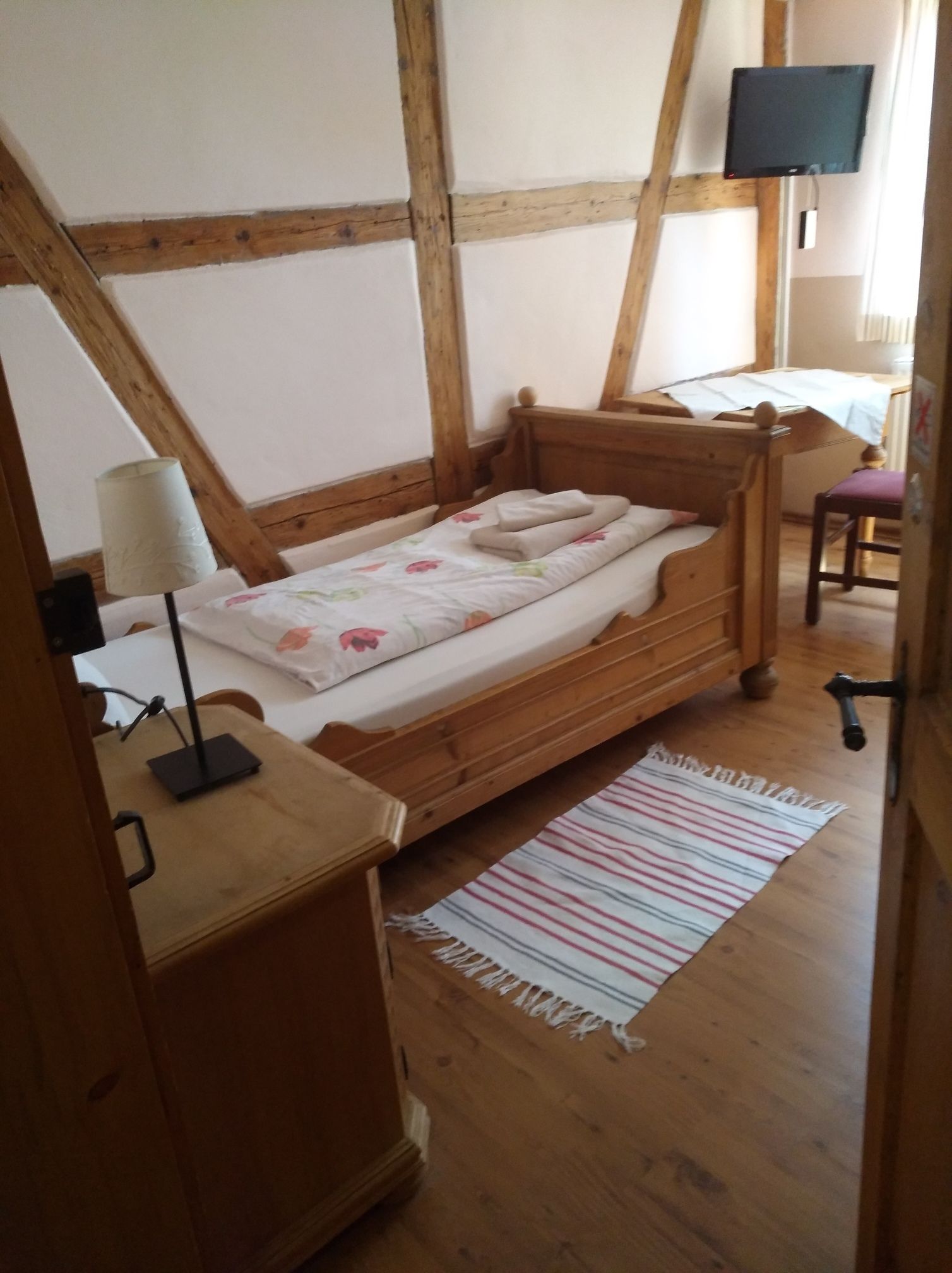 Una cama de madera en una habitación con techo inclinado, un escritorio con lámpara y un televisor en la pared.