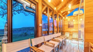 Terraza de madera con filas de sillas y mesas de lona, ventanas amplias que ofrecen vistas panorámicas y techo con vigas a la vista y lámparas colgantes.