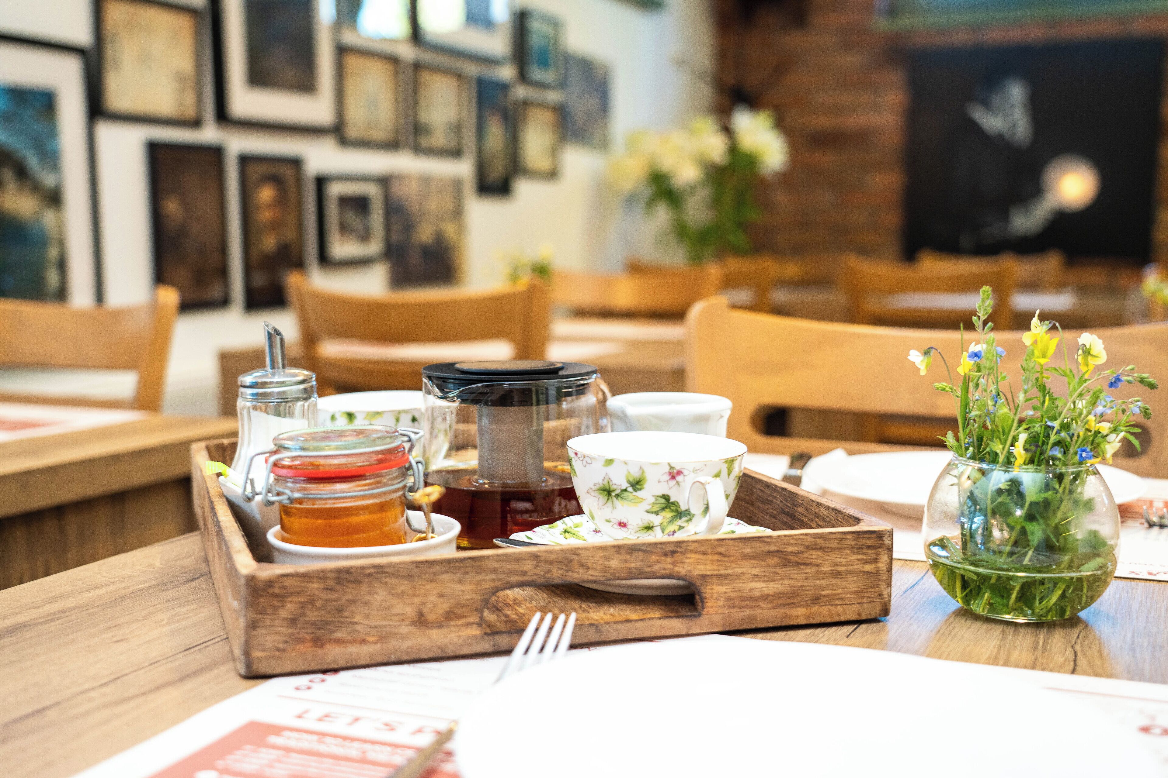 A wooden tray with a teapot, cups, and a glass jar on a table in a cafe with wooden chairs and framed pictures on the wall.