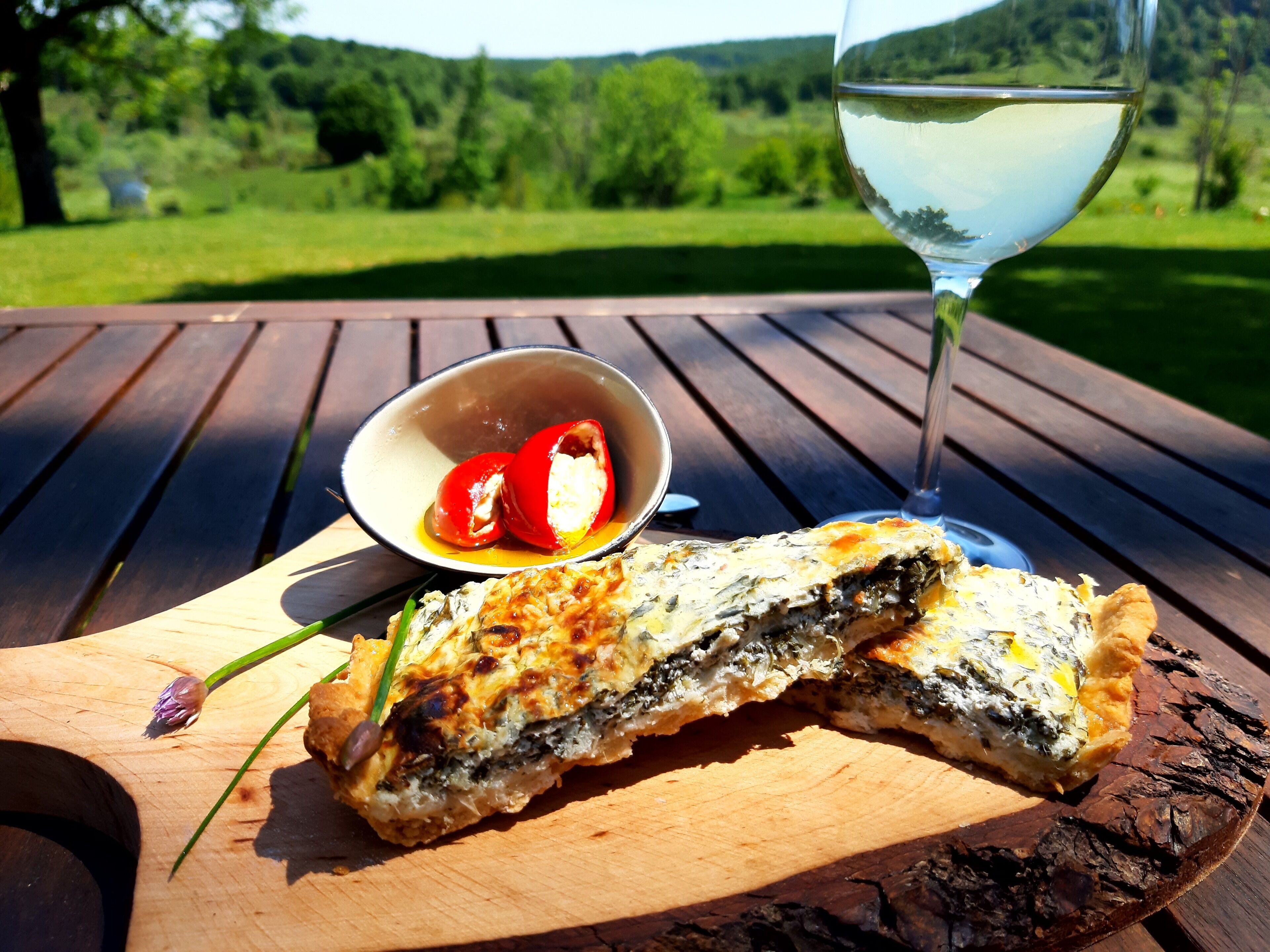 Una comida sobre una mesa de madera con una copa de vino blanco, un bol de pimientos rojos y un pastel relleno.