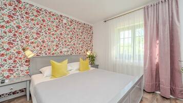 Una cama bien hecha con almohadas blancas y amarillas, una pared cubierta de papel tapiz floral, una mesita de noche con una lámpara y una ventana con cortinas traslúcidas.