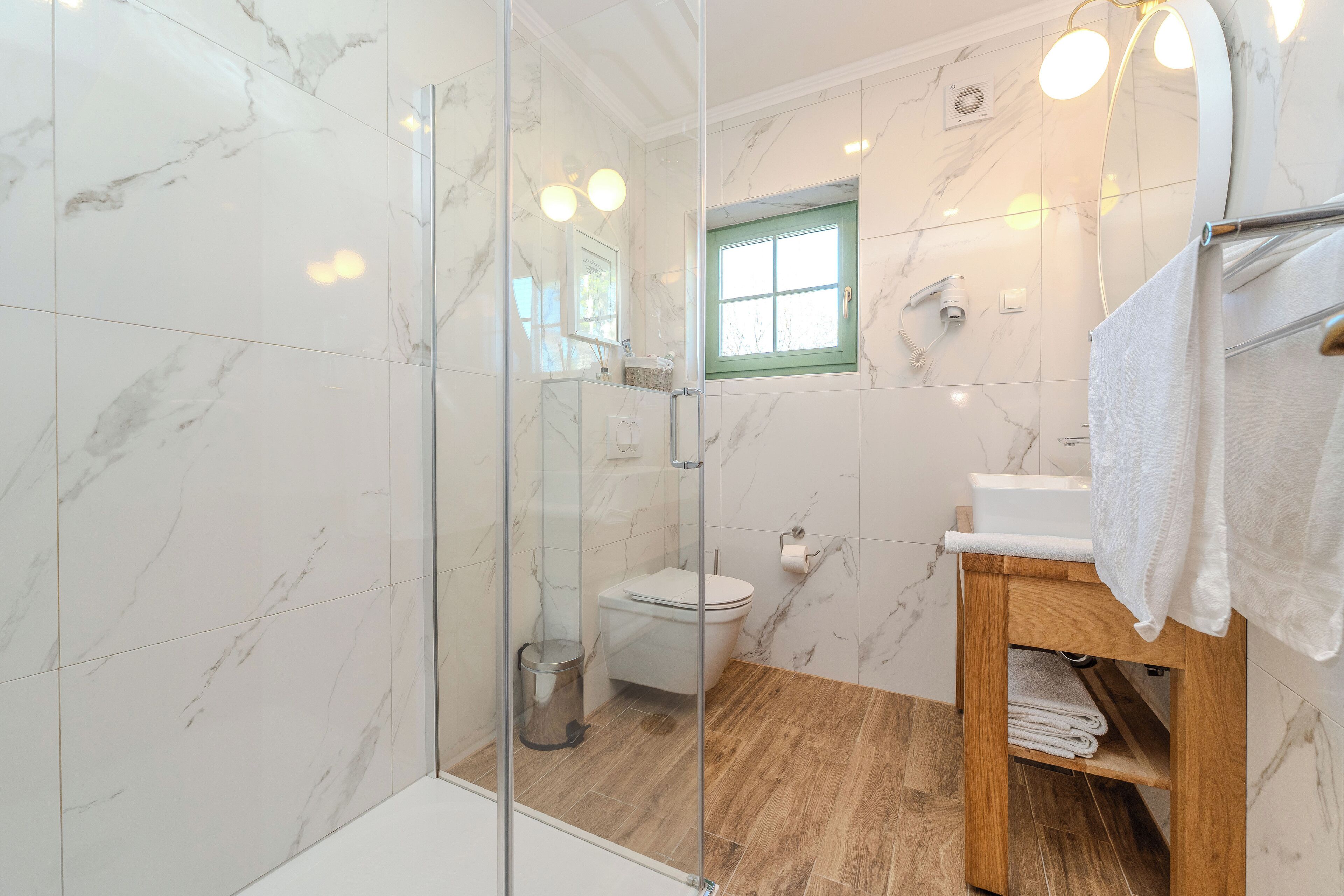 Un baño moderno con paredes de mármol, tocador de madera, cabina de ducha de vidrio y ventana con marco verde.