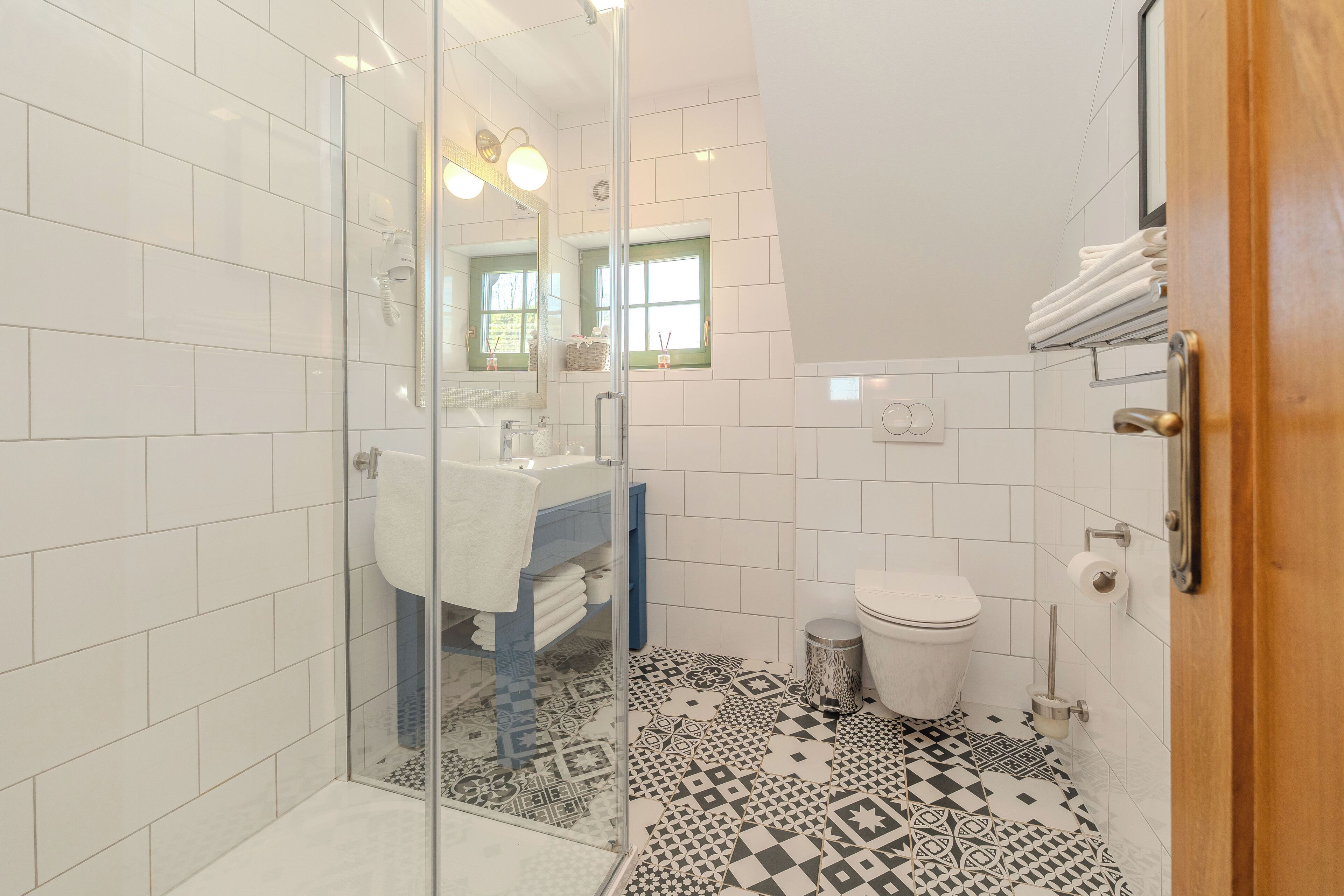 A modern bathroom with white tiles, a glass shower enclosure, a patterned floor, and a visible window with a green frame.