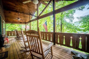 A wooden deck with rocking chairs, a flag, and a view of trees. - Alpine Rose - Pet-Friendly, Cozy Cabin w/Hot Tub (Helen)