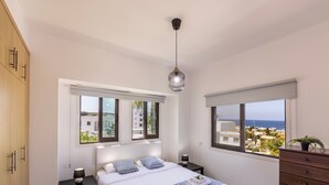 A bedroom with a bed, two bedside tables, a dresser, and two windows with views of buildings and the sea.
