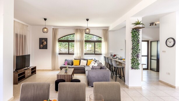 A modern living room with a sofa, coffee table, and TV. There is a dining area with chairs and a fruit basket.